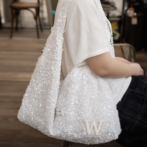 May include: A white shoulder bag covered in small, reflective sequins. The bag features a single wide strap and a prominent, embroidered capital letter W on the front lower section. A small rectangular fabric label is attached near the strap base.