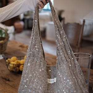 May include: A beige knit hobo bag featuring a loose, open-weave texture embellished with small, reflective silver sequins throughout the fabric. The bag includes a single integrated shoulder strap and a small white rectangular label with text.