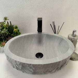 Op de afbeelding: Een grijze, ovale stenen wastafel met een zwarte kraan. De wastafel heeft een ruwe, getextureerde buitenkant en een gladde binnenkant. Een zwarte afvoer en een zeepdispenser zijn ook zichtbaar.