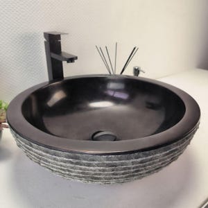 Op de afbeelding: Een donkergrijze, ronde badkamer wastafel met een getextureerde stenen basis. De wastafel heeft een glad, donker interieur en een bijpassende zwarte kraan. Een kleine zeepdispenser en een rietdiffuser zijn zichtbaar.