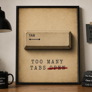 May include: A framed print with a large, textured "TAB" key and the text "TOO MANY TABS OPEN" with a red line through "OPEN". A black coffee mug with "coffee > code > repeat" is on a wooden surface. A black desk lamp and a small plant are also visible.