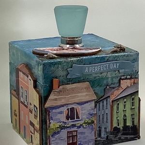 May include: A blue and green wooden box with a light blue glass knob on the top. The box is decorated with a collage of images of houses and the text "A Perfect Day".