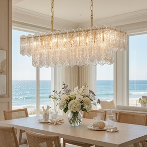 May include: A rectangular chandelier featuring multiple tiers of clear, textured glass panels suspended by gold-toned metal chains. The light fixture emits a warm glow, illuminating the intricate, scalloped edges of the glass pieces.