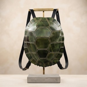 Op de afbeelding: Een groene rugzak in de vorm van een schildpadschild met een zeshoekig patroon en zichtbare lichtgekleurde stiksels. De tas heeft twee zwarte verstelbare schouderbanden die aan de boven- en zijkanten zijn bevestigd en hangt aan een metalen standaard.