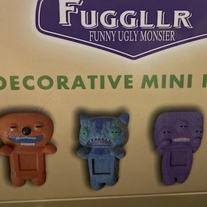 Puede incluir: Una caja de mini figuras decorativas "Fuggler". Las figuras son de varios colores, incluyendo naranja, azul, morado y amarillo. Cada figura tiene un diseño único y peculiar con rasgos y expresiones exageradas. La caja tiene el texto "Attach Me!"