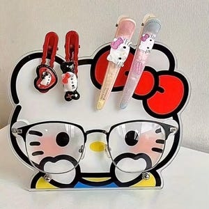 Puede incluir: Un organizador transparente con temática de Hello Kitty con gafas con montura negra, pinzas para el pelo rojas y dos limas de uñas. El organizador presenta un fondo blanco con contornos negros y detalles en rojo, y una base amarilla y azul.