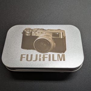 May include: A silver rectangular tin with a Fujifilm camera design etched on the lid. The camera image is centered above the word "FUJIFILM" in a bold font. The tin sits on a dark surface.
