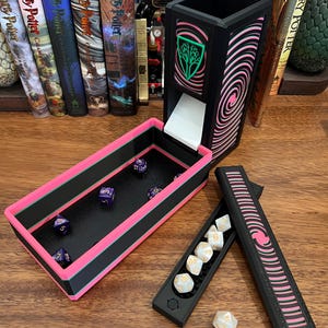 May include: A black and pink 3D-printed dice tower with a matching rectangular dice tray. The tower features a green tree emblem and a pink spiral pattern. Several purple dice are inside the tray, while a separate black case holds white dice.