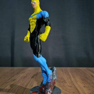 Puede incluir: Figura impresa en 3D de un superhéroe en amarillo, negro y azul. La figura está en una pose dinámica, con una base gris con salpicaduras rojas. La figura está sobre una superficie de madera. El diseño sugiere un tema de cómic o superhéroe.