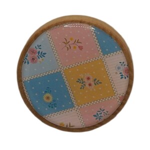 K&ouml;nnte beinhalten: Ein runder Holzuntersetzer mit Patchwork-Design. Das Patchwork zeigt Quadrate aus rosa, blauen und gelben Stoffen mit floralen Mustern. Der Untersetzer hat einen hellbraunen Holzrand.