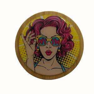 May include: Round wooden coaster featuring a colourful pop art design of a woman wearing sunglasses with the word "WOW" printed on them. The background is yellow with black polka dots.