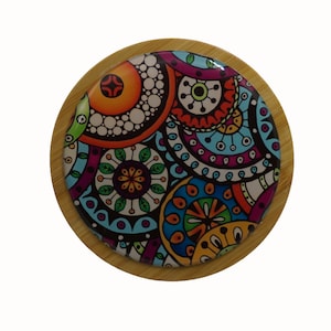 May include: A round, wooden coaster with a colourful, abstract design featuring circles, swirls, and floral patterns in shades of blue, green, orange, pink, and purple.