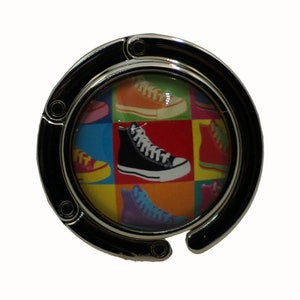 Puede incluir: Un gancho para bolso de metal plateado con un diseño redondo y colorido que presenta una variedad de zapatillas. El diseño incluye una zapatilla negra de caña alta con cordones blancos.