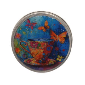 May include: A circular metal tin with a vibrant, artistic design. The design showcases a teacup and saucer adorned with floral patterns, set against a blue backdrop with butterflies. The teacup is orange, red, and yellow.