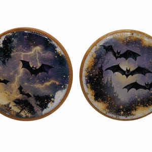 May include: Two round wooden coasters with a Halloween theme. One coaster features bats flying in front of a stormy sky with lightning. The other coaster shows bats flying in front of a castle.