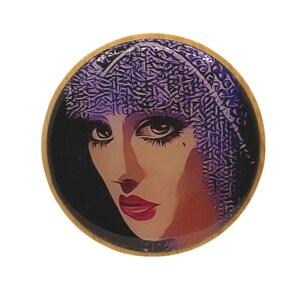 Peut inclure: Un dessous de verre rond en bois avec une image colorée d'une femme portant un couvre-chef à motifs violets et noirs. La femme a les cheveux noirs et regarde vers le bas avec une expression sérieuse.