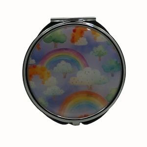 Puede incluir: Un espejo compacto plateado con un diseño colorido que presenta arcoíris, nubes y árboles sobre un fondo morado.