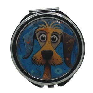 Puede incluir: Un espejo de bolsillo plateado con una imagen redonda y colorida de la cara de un perro. El perro tiene pelo marrón y amarillo, grandes ojos marrones y una nariz negra. El fondo es azul con remolinos y puntos.