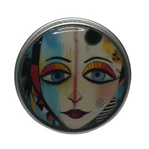 May include: A round, silver-colored pin with a colorful, abstract face design. The face has blue eyes, a red mouth, and a yellow and orange design around the eyes.