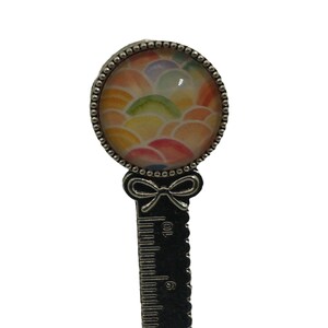 Op de afbeelding: Een zilveren bladwijzer met een regenboogkleurige glazen cabochon en een strik. De bladwijzer heeft een liniaal aan de achterkant.