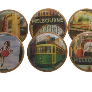May include: Six round wooden knobs with colorful images of Melbourne, Australia. The images include a tram, a building with the word "MELBOURNE" on it, a building with a dome, a building with a large mouth, and a girl jumping in the air.
