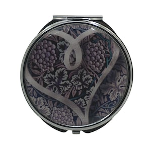 Puede incluir: Un espejo de bolsillo plateado con un diseño floral redondo. El diseño presenta una forma de corazón blanco con un patrón floral morado y azul.