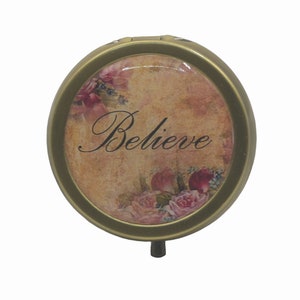 May include: A round, gold-colored trinket box with a floral design and the word "Believe" printed on it.