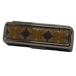 May include: A rectangular, silver-toned pill box with a hinged lid. The top features a decorative floral pattern in shades of yellow, blue, and white. The box is closed and appears to be made of metal.