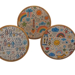 May include: Three round wooden coasters with colorful hand-drawn designs. The designs include rainbows, stars, suns, clouds, and stick figures. One coaster has the word "FAMILY" written on it.