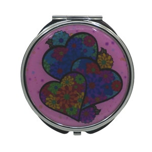 Puede incluir: Un espejo de bolsillo plateado con fondo rosa y dos corazones superpuestos decorados con flores de colores.