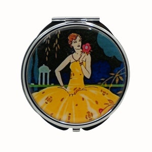 Puede incluir: Un espejo compacto plateado con una ilustración vintage de una mujer con un vestido amarillo sosteniendo una flor rosa. El fondo es un paisaje azul oscuro y verde.