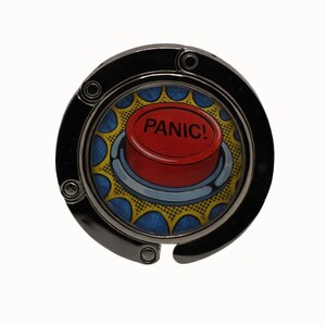Puede incluir: Un gancho para bolso de metal negro con un diseño circular. El centro presenta un botón rojo con la palabra "PANIC!" en texto blanco, sobre un fondo de estilo cómic azul y amarillo.