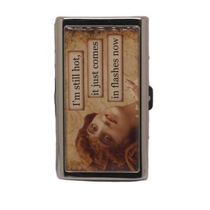 May include: A silver metal cigarette case with a rectangular design. The case features a vintage image of a woman and text that reads, "I'm still hot, it just comes in flashes now."