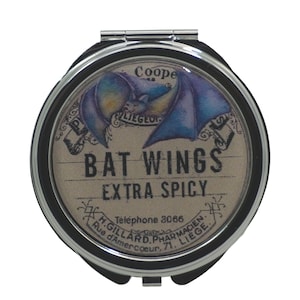 Puede incluir: Un espejo de bolsillo plateado con una tapa redonda de color crema que presenta una ilustración de estilo vintage de un murciélago con las alas extendidas. El texto "BAT WINGS EXTRA SPICY" está impreso en negro en la tapa, junto con las palabras "Teléfono 3066" y "H. GILLARD. PHARMACIEN. Rue d'Amercœur. 71. LIEGE."