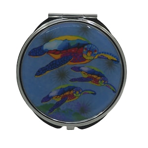 Puede incluir: Un espejo compacto plateado con un fondo azul y una imagen colorida de tres tortugas marinas nadando. Las tortugas están pintadas en tonos de azul, verde, amarillo, rojo y morado.
