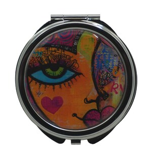 Puede incluir: Un espejo compacto plateado con un diseño abstracto colorido que presenta una cara estilizada con un gran ojo azul, labios rosados y un corazón rojo.