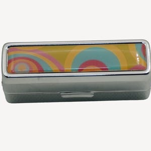 May include: A rectangular, silver-colored pill box with a colorful, retro-style design on the lid. The lid features a pattern of concentric circles in shades of yellow, teal, pink, and orange. The box is closed.
