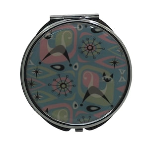 Puede incluir: Un espejo de bolsillo plateado con un diseño moderno de mediados de siglo azul, rosa y verde con gatos estilizados y estrellas.
