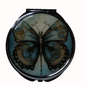 Puede incluir: Un espejo de bolsillo con un marco de metal plateado y un diseño redondo de mariposa azul y marrón con engranajes y ruedas dentadas. La mariposa tiene un cuerpo largo y delgado y alas delicadas e intrincadas.