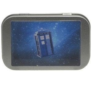 May include: A silver metal tin with a blue and white image of the TARDIS, the time machine from the Doctor Who television series, against a starry night sky background.