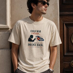 Può includere: T-shirt beige con la scritta "COLD WAR CALLED... it wants its DRAMA BACK." La grafica include una silhouette che indica una bandiera cubana e la scritta "USA * CUBA."