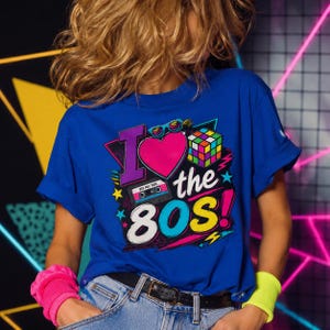 Può includere: T-shirt blu reale con una grafica colorata che dice "I love the 80s!". Il design include un cuore rosa, un cubo di Rubik, occhiali da sole e una musicassetta. La maglietta ha le maniche arrotolate.