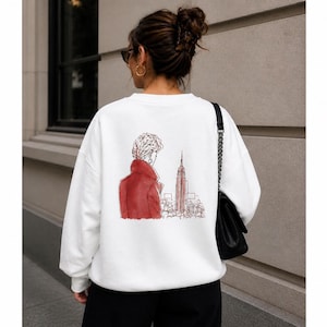 Può includere: Felpa bianca con illustrazione rossa e nera sul retro. Il design raffigura una persona con un cappotto rosso che guarda un paesaggio urbano con un edificio alto. È visibile una borsa a tracolla nera.