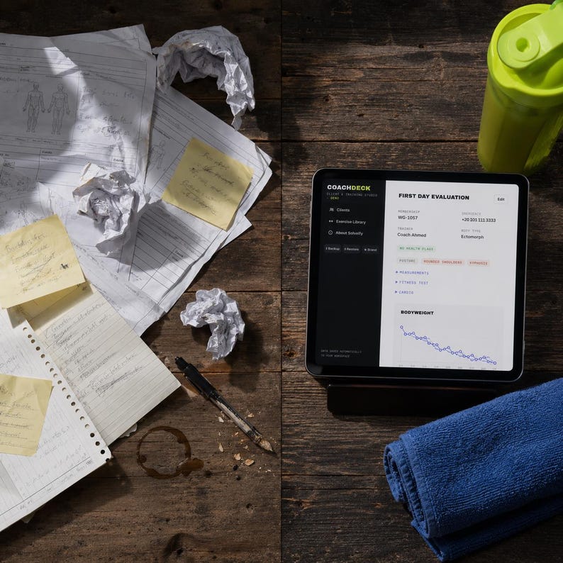 以下が含まれることがあります： CoachDeckアプリのFirst Day Evaluation画面を表示したタブレット。周囲には散らばった書類、丸めたメモ、ペン、ライムグリーンのシェイカーボトル、折りたたまれた青いタオルが置かれています。画面にはCoach-Ahmedのデータと体重が表示されています。