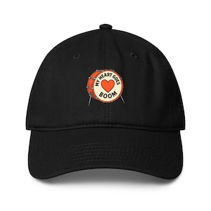 Op de afbeelding: Zwarte baseballpet met een gebogen klep. De pet heeft een geborduurd ontwerp van een trommel met een hart en de woorden "My Heart Goes Boom" in een cirkelvormig patroon. De trommel is oranje en wit met zilveren drumstokken.