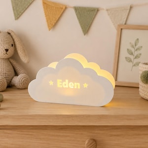Op de afbeelding: Een witte, wolkvormige lamp met een warme interne gloed, voorzien van de naam Eden in dikke letters geflankeerd door twee kleine sterren. De lamp is afgewerkt met een gladde, matte laag en verspreidt een zacht licht vanuit het midden.