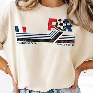 K&ouml;nnte beinhalten: Cremefarbenes T-Shirt mit einem Used-Look-Grafikdesign. Das Design zeigt die Buchstaben "FR" in Rot und Blau, einen Fu&szlig;ball und den Text "FRANCE SOCCER WORLD CUP '26". Das Shirt hat einen Vintage-Look.