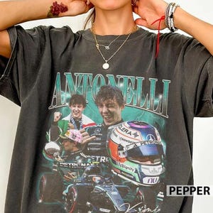 May include: A charcoal gray short-sleeve t-shirt featuring a graphic print of a race car driver, a racing helmet, and a Formula 1 car. The text ANTONELLI is printed in large teal letters above the imagery, which includes a green lightning effect.