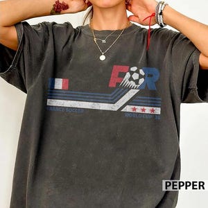 K&ouml;nnte beinhalten: Dunkelgraues T-Shirt mit einem grafischen Design, das die franz&ouml;sische Flagge, einen Fu&szlig;ball und den Text "FOR" in Rot und Blau zeigt. Das Shirt enth&auml;lt auch die W&ouml;rter "FRANCE SOCCER" und "WORLD CUP 26".