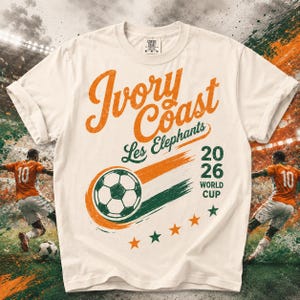 May include: A cream-colored t-shirt featuring the text Ivory Coast Les Elephants and 2026 World Cup. The design includes a soccer ball graphic with orange and green brush strokes, five small stars, and two soccer players in orange jerseys.